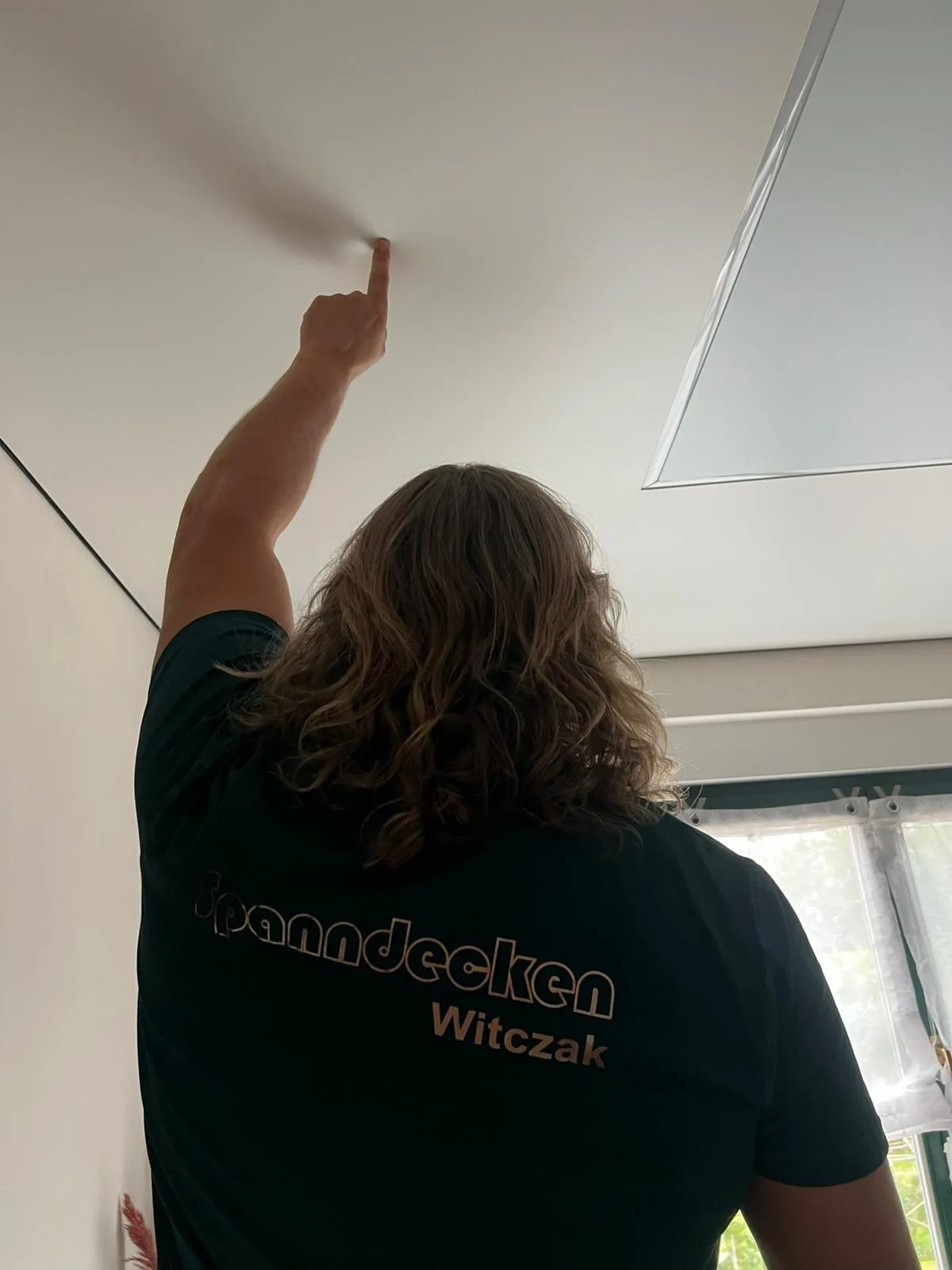 Mike Witczak im Firmenshirt, zeigt auf die fertige Spanndecke